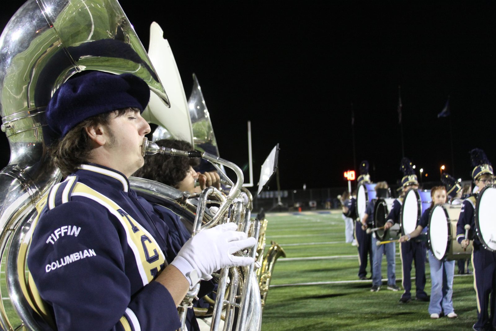 A Columbian band member plays a sousaphone on the field at night as the drumline stands nearby.