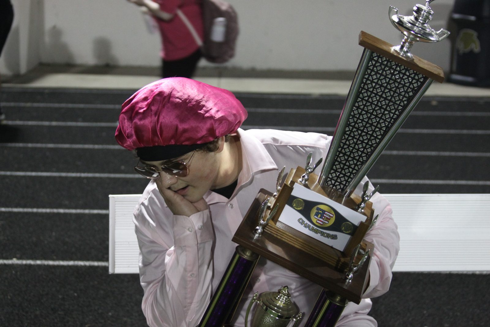 A Columbian student in a pink shirt, pink cap and sunglasses poses with a large championship trophy.