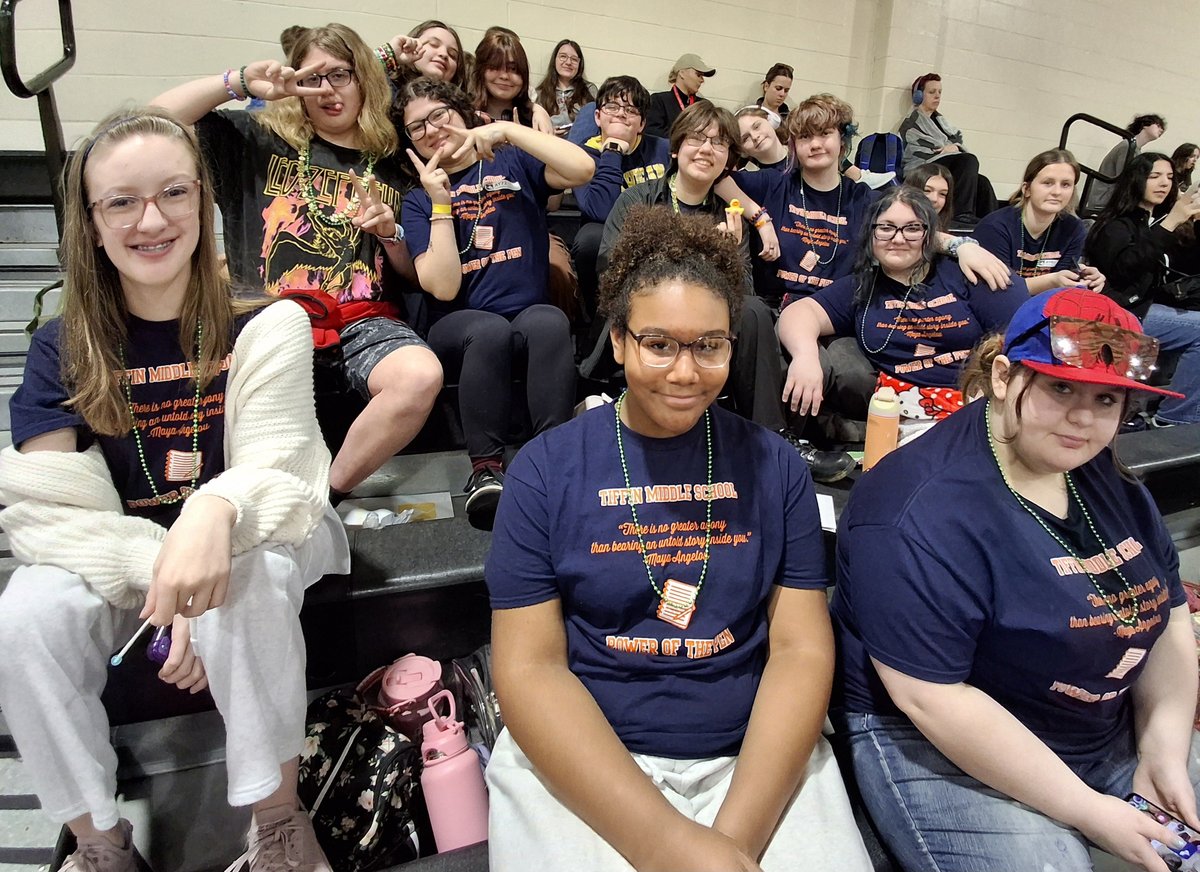 Tiffin Middle School students wearing navy Power of the Pen shirts and green bead necklaces smile from gym bleachers.