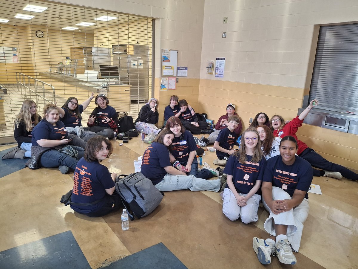 A group of Tiffin Middle School students in navy Power of the Pen shirts sit together on the floor of a school commons area.